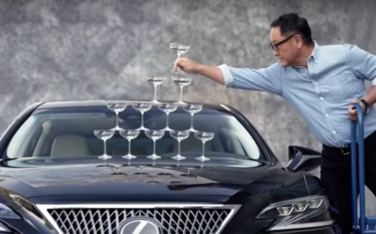 Akio Toyoda builds a tower of champagne glasses on the hood of a Lexus LS 500